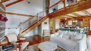Amplia sala de estar con una escalera de madera, un sofá blanco y una cocina al fondo.
