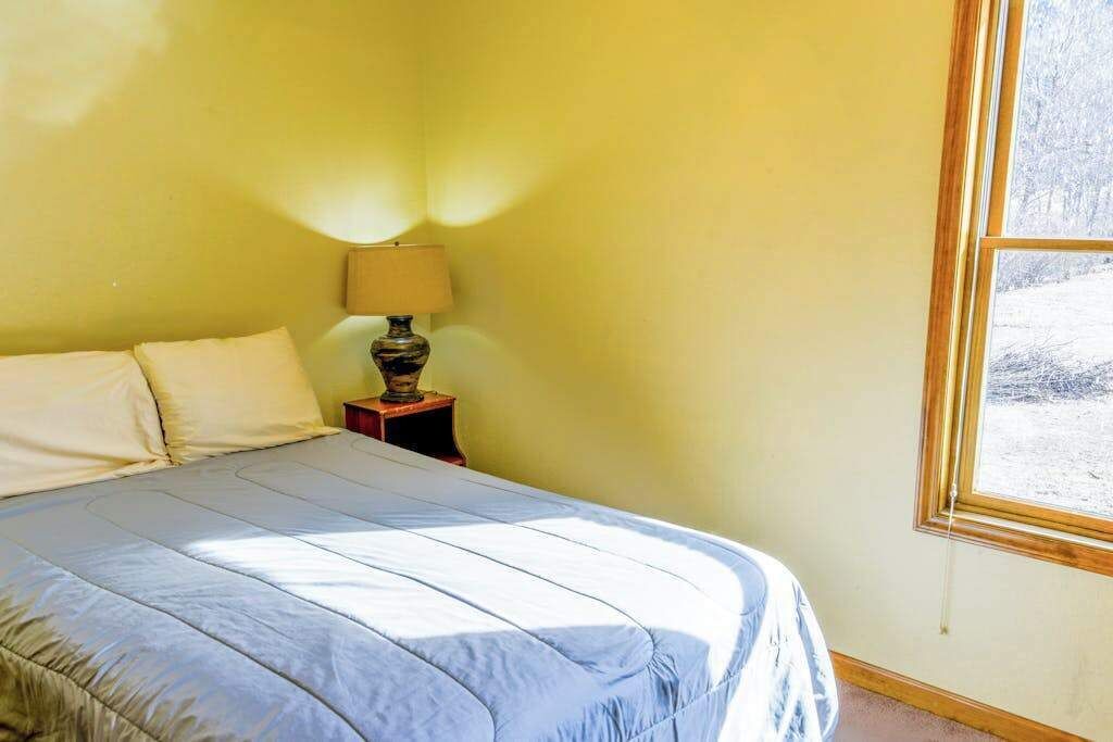 Ein Zimmer mit einem Bett, einem Nachttisch mit Lampe und einem Fenster mit Blick auf Bäume.