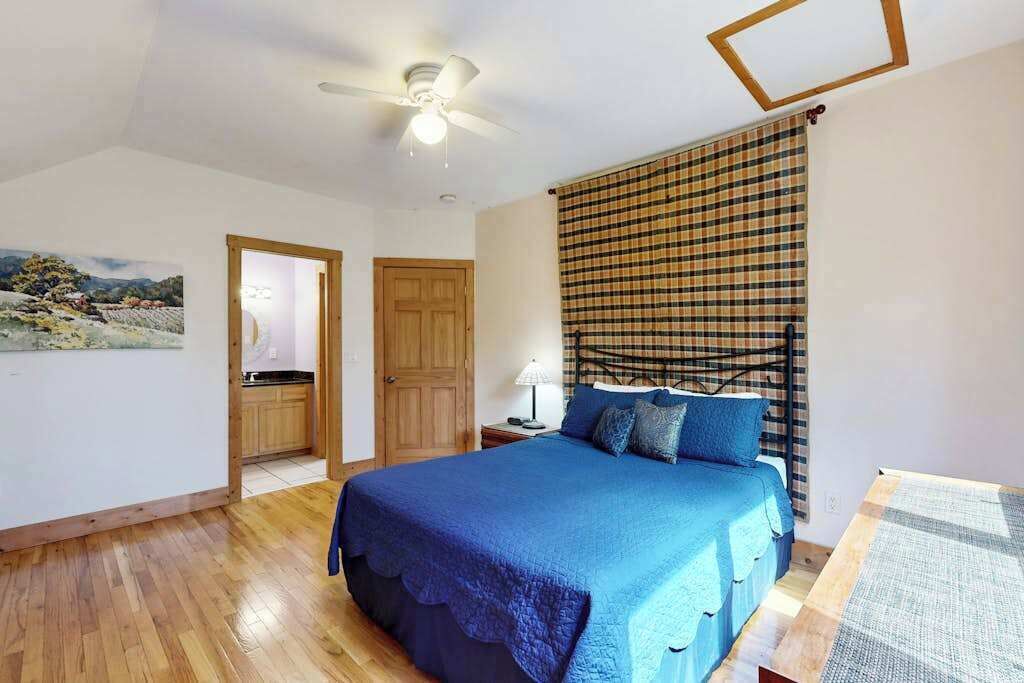 Un dormitorio con cama, ventilador de techo, ventana con persiana a cuadros y puerta que da al baño.