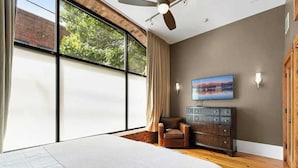 Een slaapkamer met een groot raam, een plafondventilator, een bed, een stoel, een dressoir en een aan de muur bevestigde televisie.