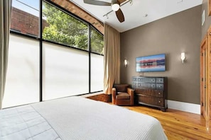 Ein Schlafzimmer mit großem Fenster, Deckenventilator, Bett, Sessel, Kommode und Wandfernseher.