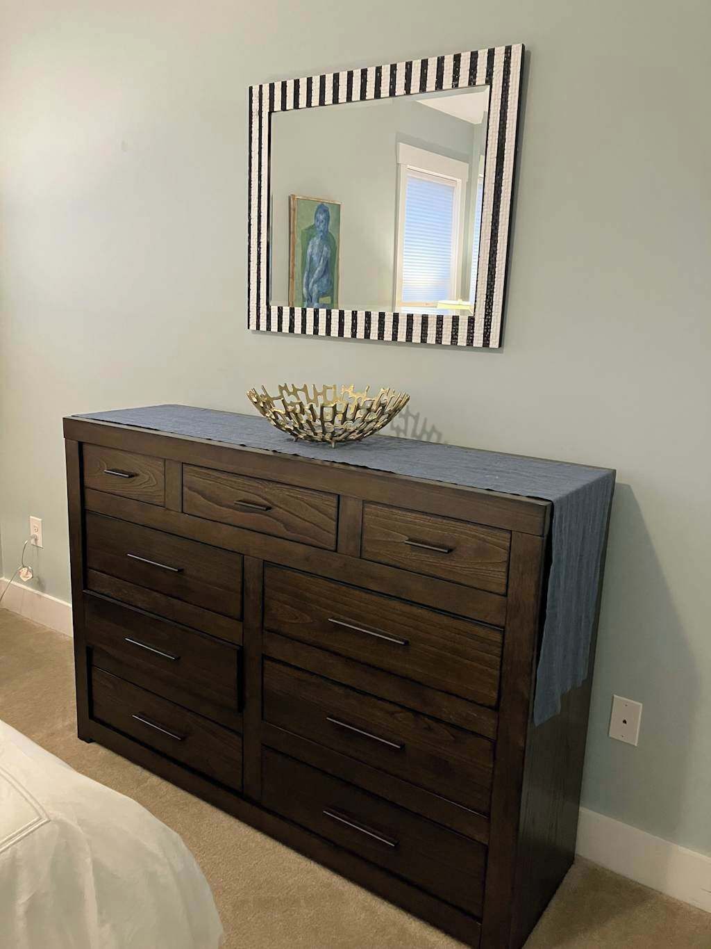 Un aparador de madera oscura con varios cajones y un cuenco decorativo encima, frente a una pared clara con un espejo a rayas en la parte superior.