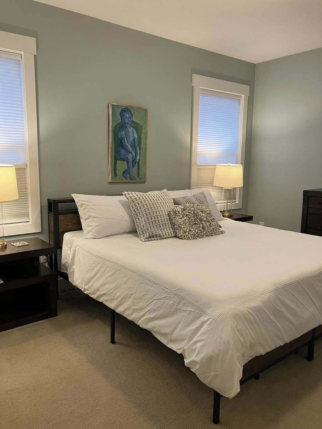 Une chambre à coucher avec un lit, deux tables de chevet, un tableau et deux fenêtres équipées de stores.