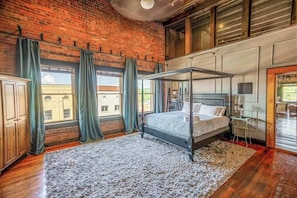 A bedroom with a four-poster bed, wooden flooring, a brick wall, and large windows with curtains.