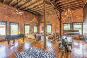 A spacious room with exposed brick walls, a grand piano, a dining area with a chandelier, and large windows.