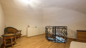 Une pièce avec un plancher en bois, une armoire blanche, une chaise en osier et un cadre de lit en fer forgé.