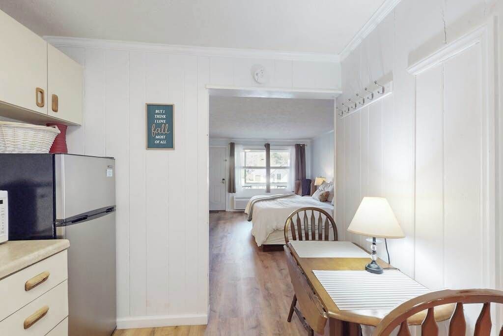 Cocina compacta con refrigerador, mesa de comedor con sillas y una cama en la habitación contigua.