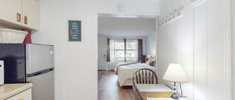 Cocina compacta con refrigerador, mesa de comedor con sillas y una cama en la habitación contigua.