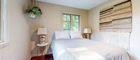 A bedroom with a bed, two bedside tables, a hanging plant, a window, and a wall tapestry.