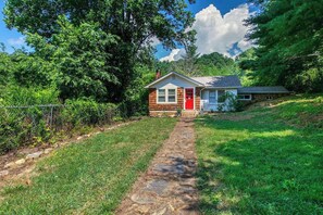A small house with a stone pathway leading to the front door, surrounded by lush greenery and trees. - Well-appointed home with a gas grill and fire table (Candler)