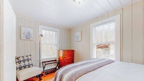 Une chambre à coucher comprenant un lit, une commode, un banc et deux fenêtres équipées de stores.
