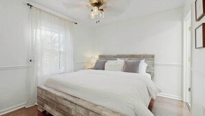 Een slaapkamer met een houten hoofdbord, een bed met witte beddengoed, een plafondventilator en een raam met dunne gordijnen.