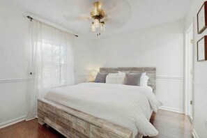 A bedroom with a wooden headboard, a bed with white bedding, a ceiling fan, and a window with sheer curtains. - Charming 4BR retreat with Full Kitchen, Wifi and Patio | Pet-friendly (Arden)