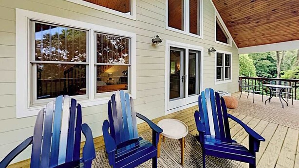 Un porche cubierto con sillas Adirondack y una mesa pequeña, terraza de madera y una casa con ventanas grandes.