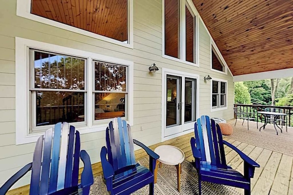 Eine überdachte Veranda mit Adirondack-Stühlen und einem kleinen Tisch, eine Holzterrasse und ein Haus mit großen Fenstern.