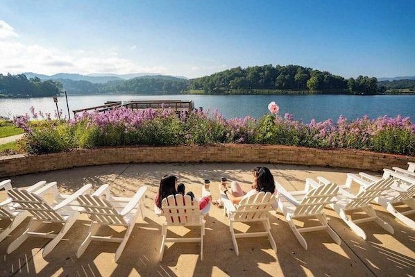 A lakeside seating area with Adirondack chairs, a wooden deck, and a view of a lake and hills.