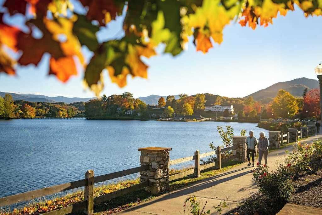 A lakeside walkway with stone railings, a stone bench, and a view of a lake and mountains.