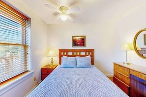 A bedroom with a bed, a ceiling fan, a nightstand with a lamp, a dresser, and a window with blinds.