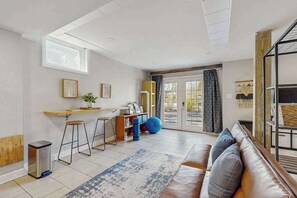 Ein modernes Wohnzimmer mit einer Couch, einem Couchtisch, einer kleinen Küchenzeile mit Barhocker und einer Glastür, die zu einem Außenbereich führt.