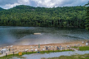 A lakeside view with a sandy beach, lounge chairs, and a forested hill in the background.