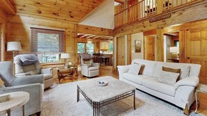 Ein geräumiges Wohnzimmer mit hoher Decke, Holzwänden, einem weißen Sofa und einem durch ein Fenster einsehbaren Essbereich.