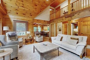 Ein geräumiges Wohnzimmer mit hoher Decke, Holzwänden, einem weißen Sofa und einem durch ein Fenster einsehbaren Essbereich.