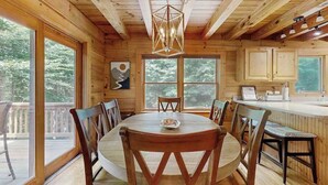 Ein Holzblockhaus-Interieur mit Esstisch, Stühlen, einer Küchenzeile und großen Fenstern.