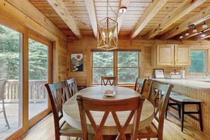 Ein Holzblockhaus-Interieur mit Esstisch, Stühlen, einer Küchenzeile und großen Fenstern.