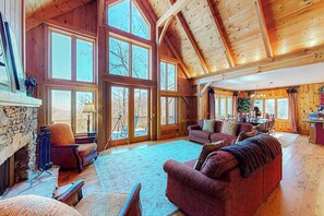 A spacious living room with a stone fireplace, large windows, and wooden beams. - Updated mountain view cabin with vaulted ceilings, large deck, & chef's kitchen (Sapphire)