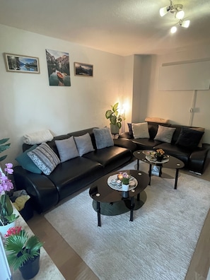 A living room with a black leather sofa, two coffee tables, and a white rug. There are three framed pictures on the wall and a potted plant on the sofa.