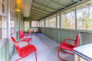 A covered porch with red metal chairs, a table, and a view of trees through large windows. - Charlotte Cabin (Waynesville)