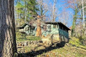 A rustic cabin with a stone foundation, wooden exterior, and a green roof, surrounded by trees and a stone pathway. - Charlotte Cabin (Waynesville)