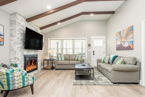A living room with a fireplace, a sofa, a coffee table, and a TV mounted on a stone wall. - Contemporary dog-friendly mountain home with firepit, grill, enclosed backyard (Swannanoa)
