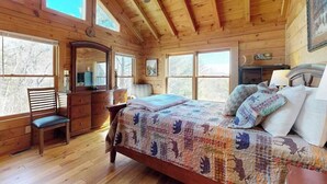 Ein Schlafzimmer in einer Holzhütte mit Bett, Schminktisch, Stuhl und großen Fenstern.