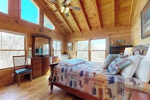 Ein Schlafzimmer in einer Holzhütte mit Bett, Schminktisch, Stuhl und großen Fenstern.