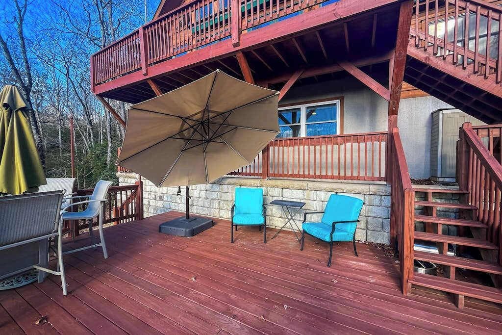 Une terrasse en bois, meublée pour l’extérieur, avec un grand parasol et un escalier menant à un deuxième niveau.