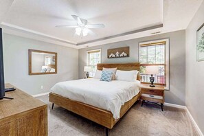 Ein Schlafzimmer mit einem Bett, Deckenventilator, Nachttisch, Lampe, Spiegel und einem Fenster mit Jalousien.
