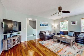 Ein Wohnzimmer mit Holzboden, einem Flachbildfernseher, einem Deckenventilator, einem Couchtisch, zwei Sofas und einem gemusterten Teppich.