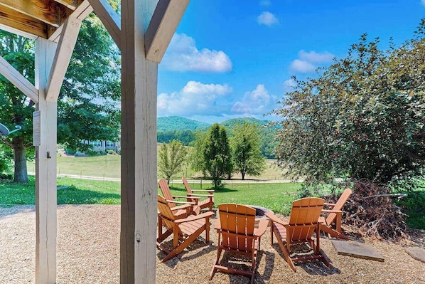Ein überdachter Sitzbereich im Freien mit Adirondack-Stühlen und einem Feuerkorb, mit Blick auf eine grüne Landschaft mit Bäumen und Hügeln.