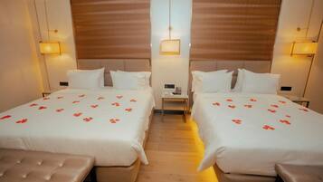 Habitación de hotel con dos camas, cada una con cubrecama blanco y motivos florales rojos, una mesita de noche con lámpara y un banco al pie de las camas.
