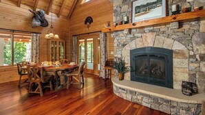 Un interior de madera con chimenea de piedra, mesa de comedor, sillas y vistas amplias a través de grandes ventanales.
