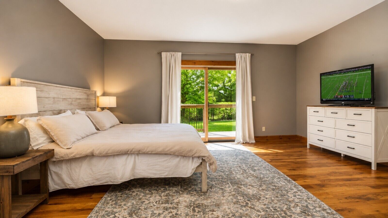 A bedroom with a bed, bedside table, lamp, TV, and a view of greenery through a window.