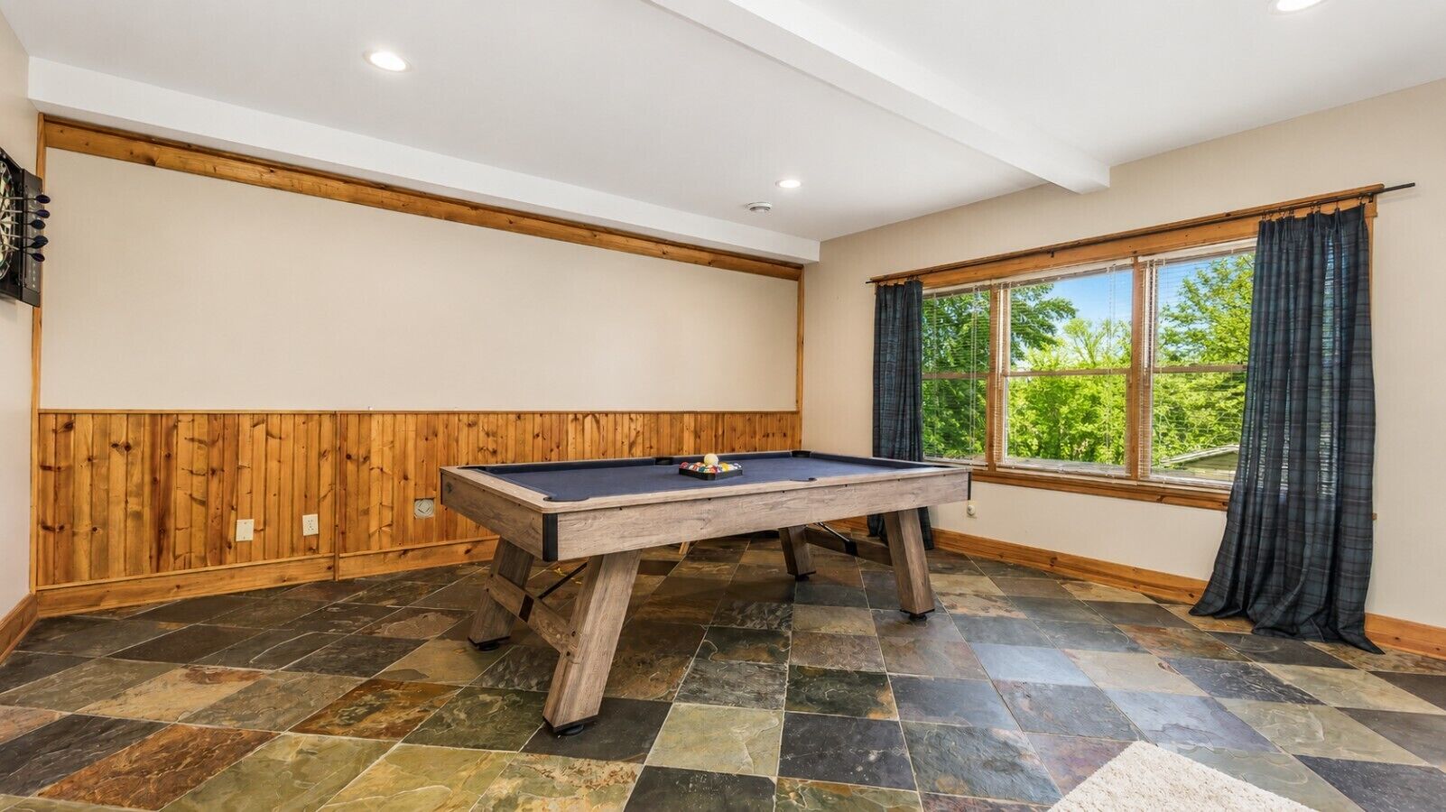 A pool table in a room with wooden paneling, a large window with curtains, and a stone tile floor.