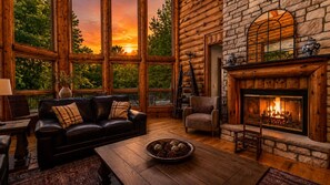 Interior de una cabaña de troncos con chimenea, sofá de cuero y amplias ventanas.