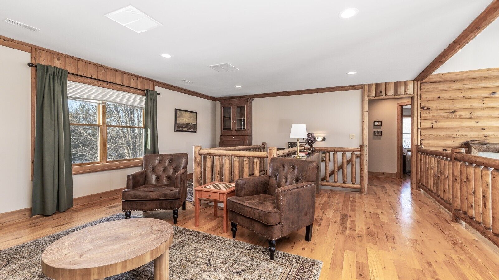 A spacious living room with wooden flooring, leather armchairs, a wooden coffee table, and a view of the outside through large windows.