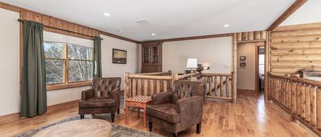 A spacious living room with wooden flooring, leather armchairs, a wooden coffee table, and a view of the outside through large windows.