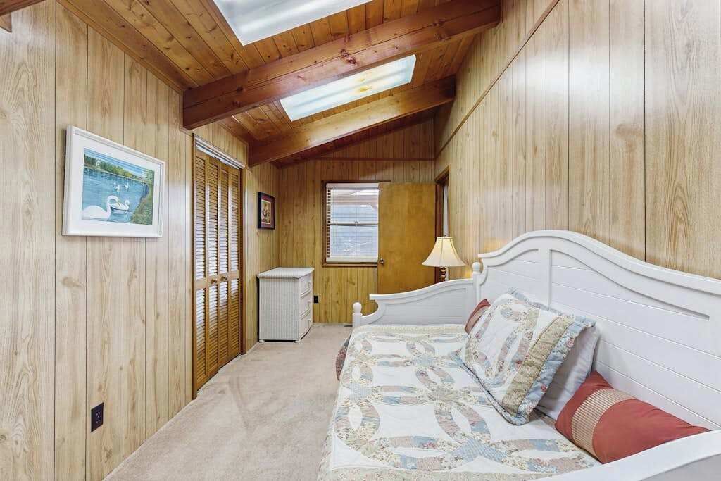 Une chambre avec des murs en bois, un lit avec une literie à motifs, une table de chevet, une lampe et une fenêtre avec des stores.