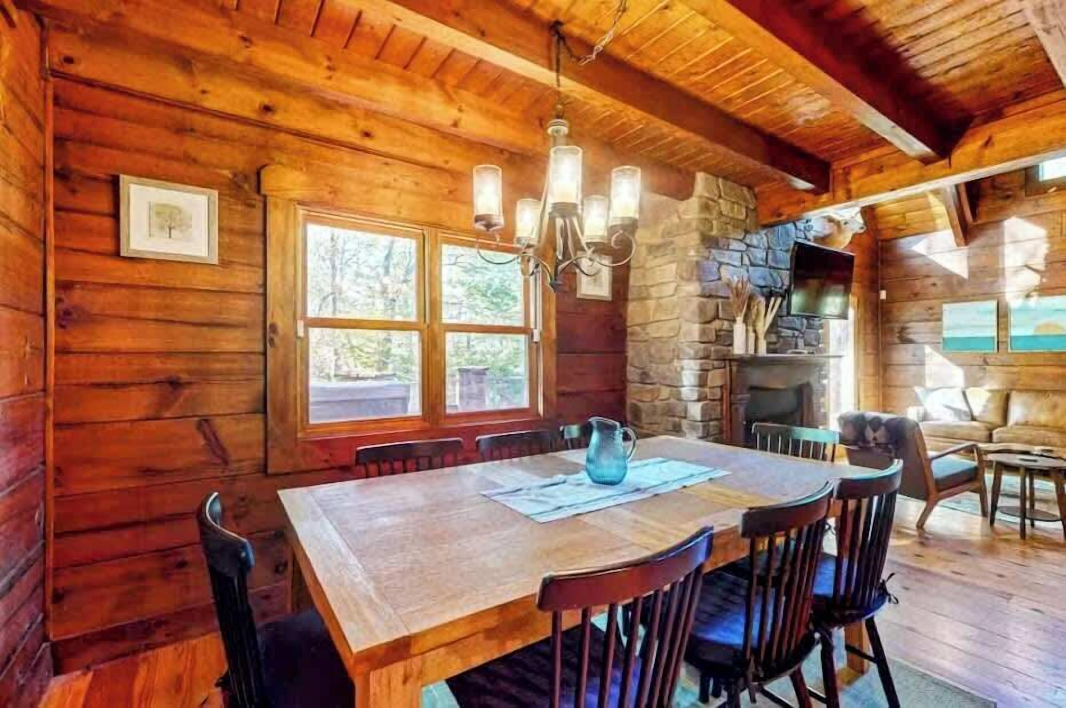 A wooden cabin interior with a dining table, chairs, a chandelier, a stone fireplace, and a large window.