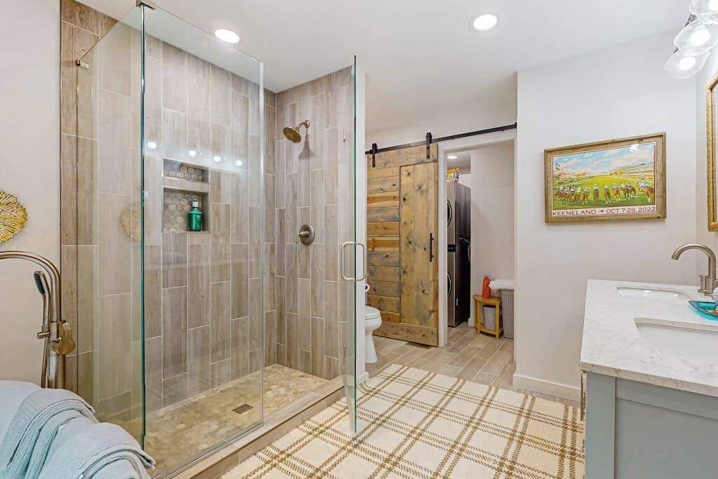 A modern bathroom with a glass-enclosed shower, a wooden door, a white sink, and a framed picture on the wall.