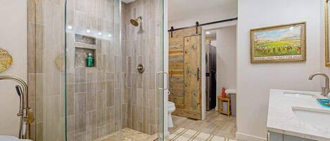 A modern bathroom with a glass-enclosed shower, a wooden door, a white sink, and a framed picture on the wall.
